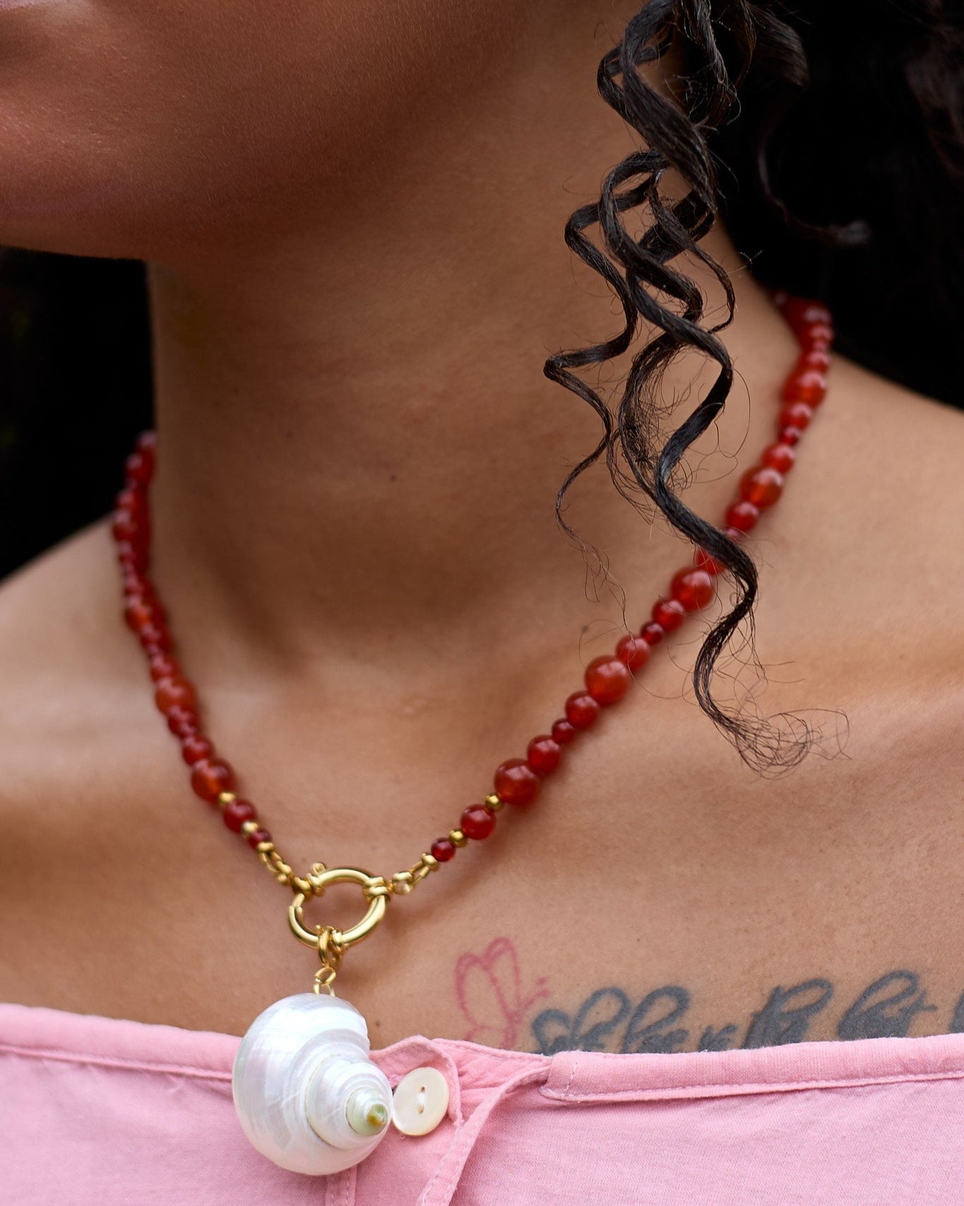 Person wearing a red beaded necklace with a large pearl pendant, pink top, and visible tattoos.
